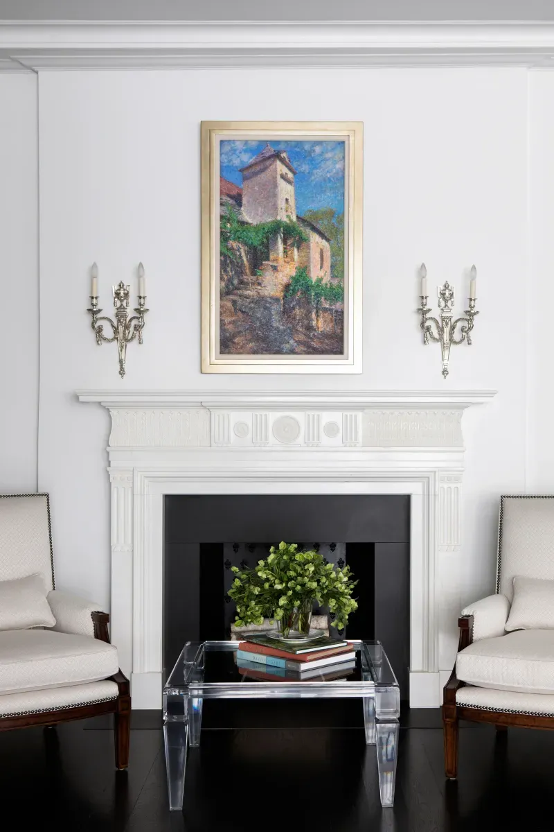 A sophisticated living room with a white fireplace adorned by a painting of a rustic landscape, flanked by two silver candelabra wall sconces. Symmetrical beige armchairs frame a clear, modern coffee table holding greenery and a stack of books on a glossy black floor.